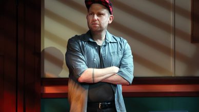 A man wearing a red checked cap and grey shirt stands with his arms crossed, looking to the side. He is indoors, lit by sunlight streaming through blinds, casting striped shadows on the wall behind him.