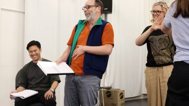 Three adults stand indoors, laughing and holding scripts. One person sits smiling with a script in hand, while the others stand, appearing to enjoy a lively conversation in a rehearsal or casual setting.
