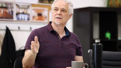 An older man with grey hair and a short beard gestures with his left hand whilst seated at a table. Papers, a mug, and a black bottle are in front of him. Blurred photos are visible on the wall behind him.