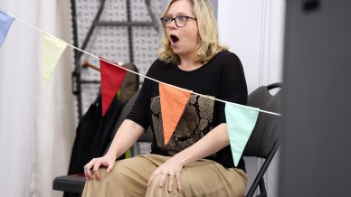 A woman with blonde hair and glasses sits on a chair, looking surprised with her mouth open. Colourful bunting hangs in front of her. She is indoors, wearing a black top and beige trousers.