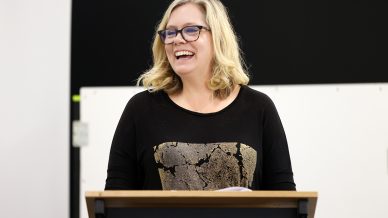 A woman with blonde hair and glasses smiles whilst standing behind a lectern, appearing to give a speech or presentation. She is wearing a black top with a gold, cracked pattern on the front.