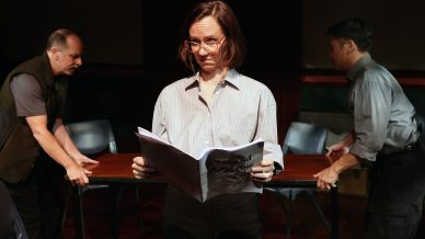 A woman with glasses stands holding a script and looking down thoughtfully, whilst two men on either side move a table behind her in a dimly lit room.