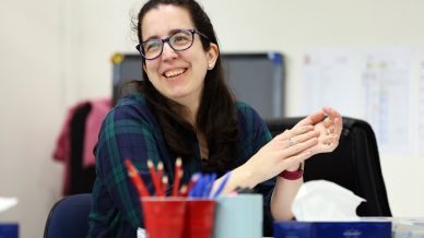 A woman with glasses and long dark hair, wearing a green and blue checked shirt, smiles whilst sitting at a desk with red pencil pots and office supplies in the foreground.