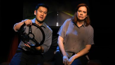 Two actors sit side by side on stage, with one holding a steering wheel as if driving. A nighttime road scene is projected behind them, suggesting they are in a car. Both have serious expressions.