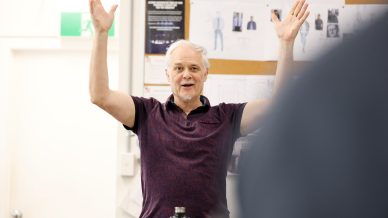 An older man with grey hair raises both arms enthusiastically while speaking in a brightly lit room with papers and sketches pinned on a noticeboard behind him.