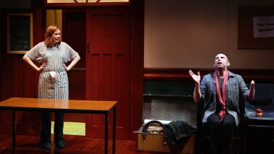 A woman in an apron stands by a table, looking at a man who is sitting with arms raised expressively. The scene takes place in a room with red doors, wooden floors, and a signboard on the wall.