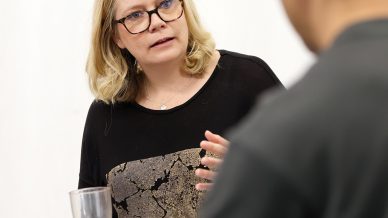 A woman with blonde hair and glasses sits holding a clear cup, speaking to a man with dark hair whose back is to the camera, against a plain white background.