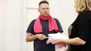 A man in a dark T-shirt and pink scarf looks intently at a woman holding papers. They appear to be in discussion, with a white background behind them.