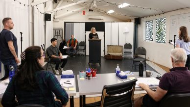 Seven people are in a rehearsal room; one person stands at a lectern speaking, while others sit or stand with scripts, watching. Tables with papers and art supplies are in the foreground. The room is bright and organised.