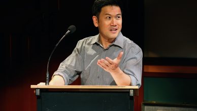 A man in a grey shirt stands behind a lectern, speaking into a microphone with one hand gesturing, appearing engaged and expressive. The background is dimly lit with dark red and green tones.