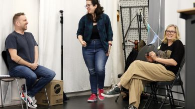 Three people are interacting in a casual indoor setting. Two are seated in folding chairs, smiling, whilst one stands in the centre, also smiling. There is a box, a microphone stand, and some equipment in the background.