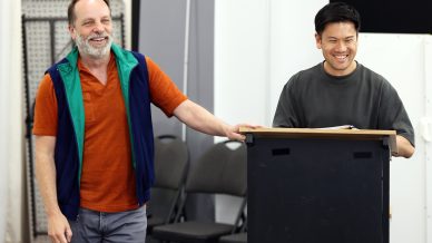 Two men stand indoors; one is smiling behind a lectern, and the other, wearing a colourful waistcoat, stands beside him also smiling. Several empty chairs are visible in the background.