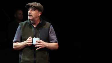 A man wearing a flat cap and a fleece gilet holds a white mug and looks to the side; another person is blurred in the dark background.