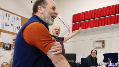 Three people in an office, two men and one woman, are smiling and laughing. One man in the foreground wears a blue waistcoat and orange shirt; another stands with arm raised. The woman sits at a desk in the background.