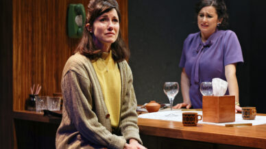 Two women in a kitchen setting look distressed. One sits on a worktop in a beige cardigan and yellow shirt, while the other stands behind the worktop in a purple top. Both appear upset; empty wine glasses and mugs are on the worktop.