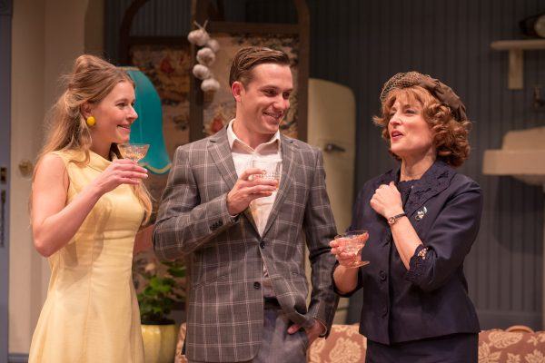 Three people dressed in vintage clothing stand indoors, holding drinks and smiling as they engage in conversation. The setting features retro decor, including hanging garlic and a patterned sofa.
