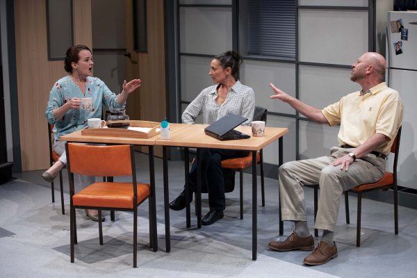 Three people sit around a square table in an office break room, engaged in conversation. One woman gestures with a mug in hand, while another woman and a man listen, seated with coffee cups and a laptop on the table.