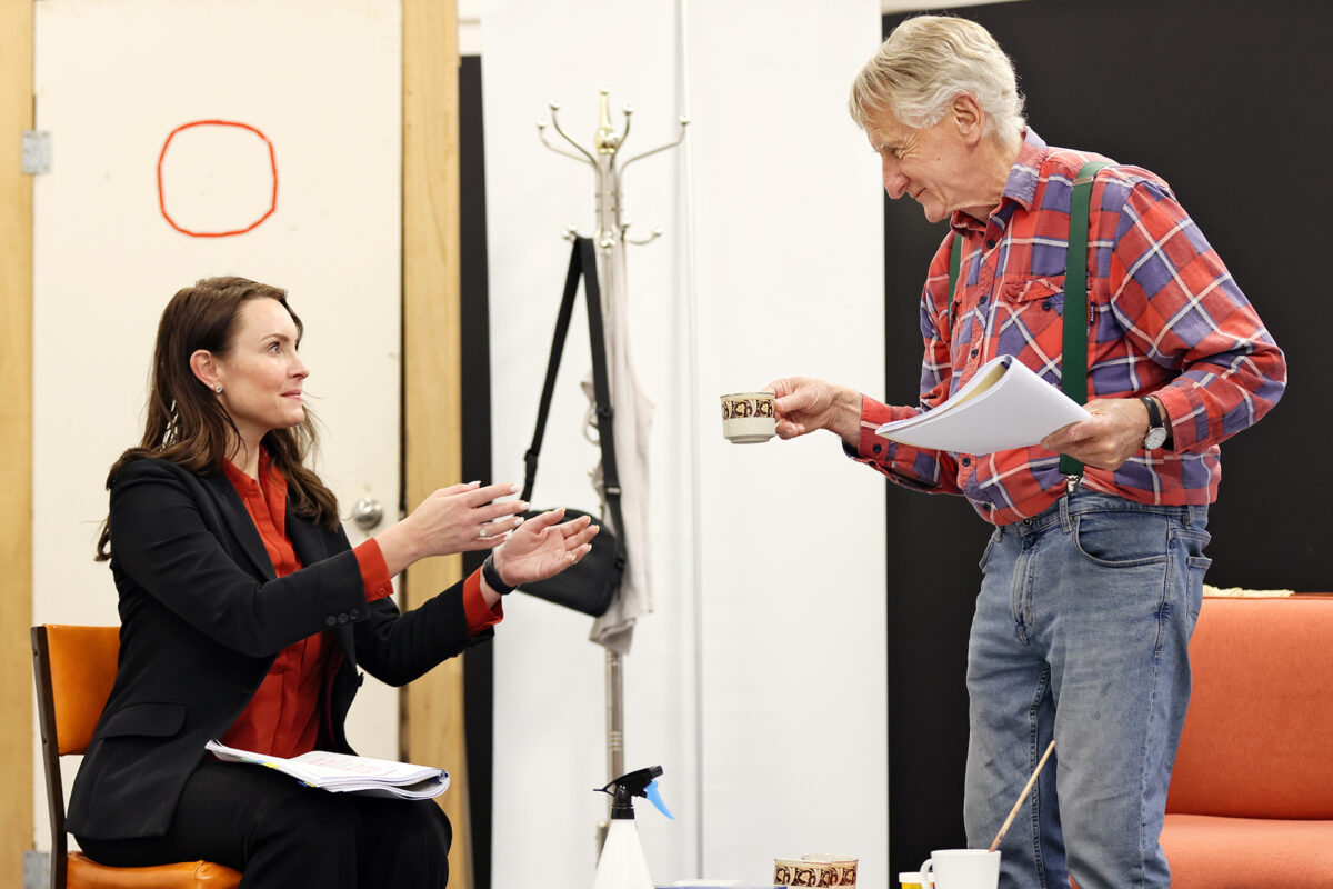 A woman in a black blazer sits holding papers and reaches for a cup of coffee offered by an older man in a checked shirt and braces. They appear to be rehearsing in a room with chairs, a coat stand, and a spray bottle.