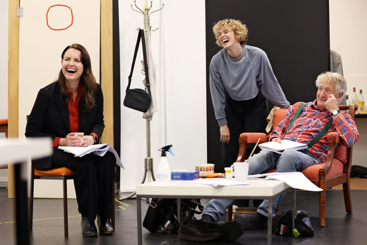Three people laugh together during a rehearsal. One woman sits in a chair holding papers, another stands leaning on a chair, and an older man sits relaxed with notes. Props and scripts are scattered around the brightly lit room.