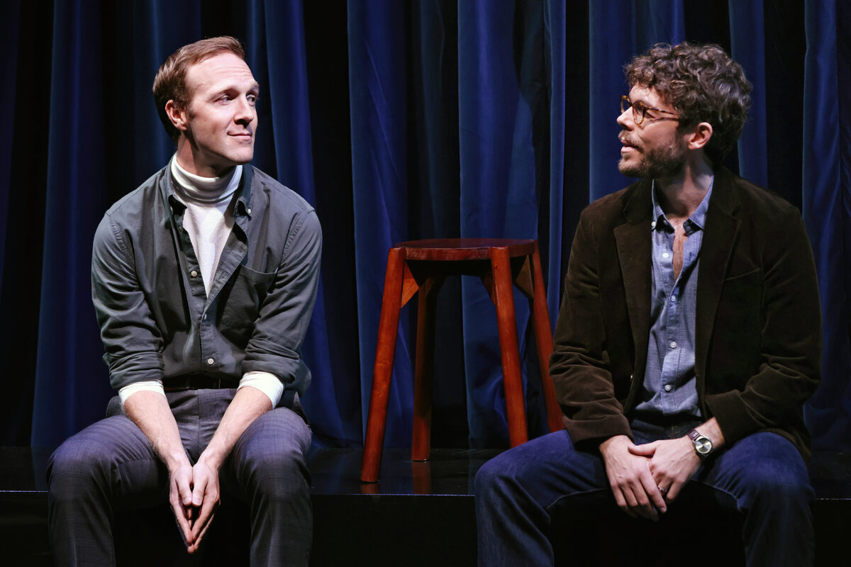 Two men sit on a stage in front of dark blue curtains, facing each other. One wears a polo neck and jacket, the other wears glasses and a brown blazer. There is a wooden stool between them in the background.