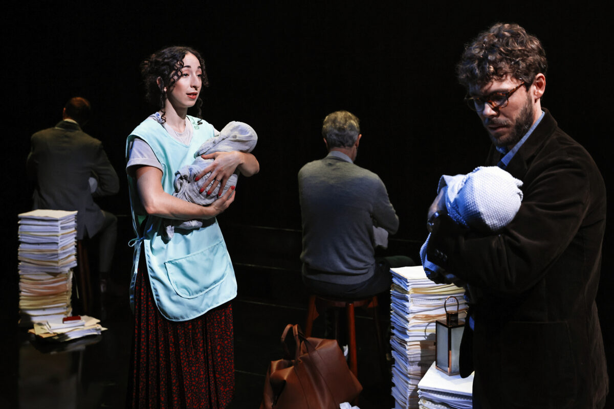 A woman and a man, each holding a baby doll, stand among piles of papers and two seated figures with their backs turned. The scene appears theatrical with dramatic lighting and dark background.