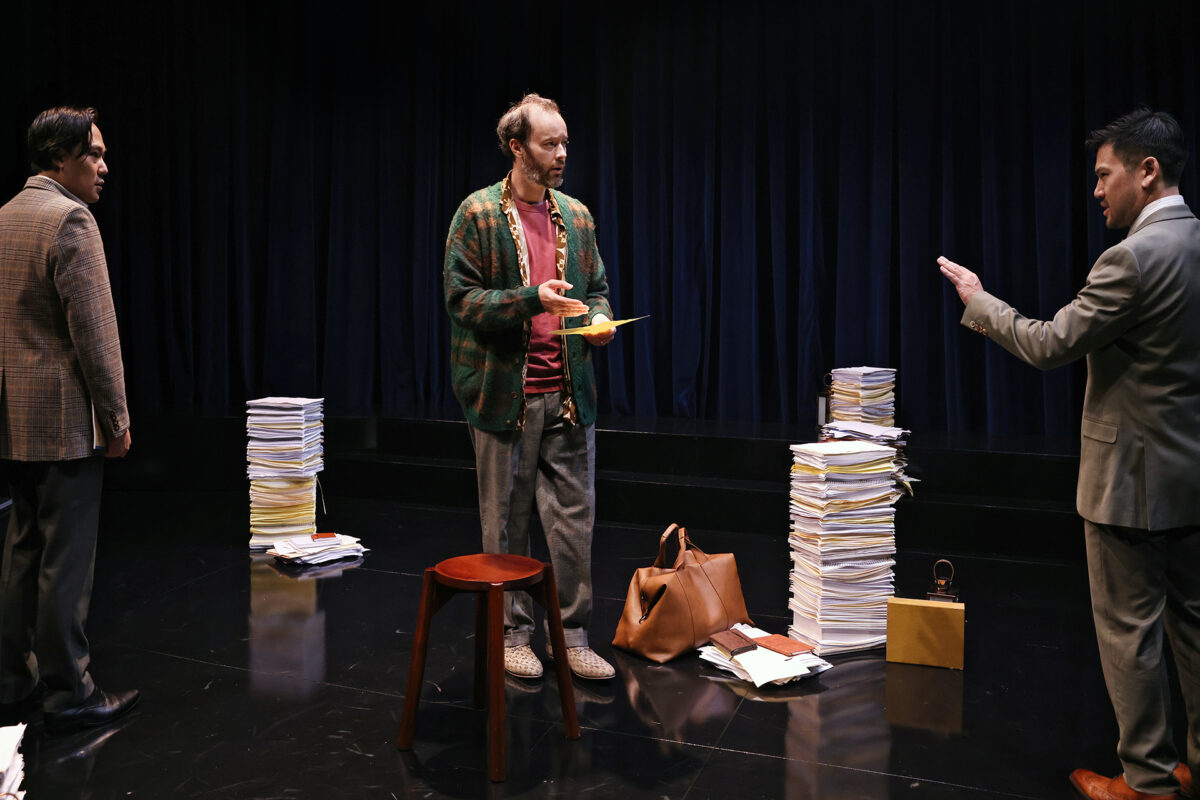 Three men stand on a stage surrounded by tall stacks of papers. One man in the centre holds papers and looks at another man who raises a hand as if gesturing or stopping him. A stool and brown bag are on the floor.