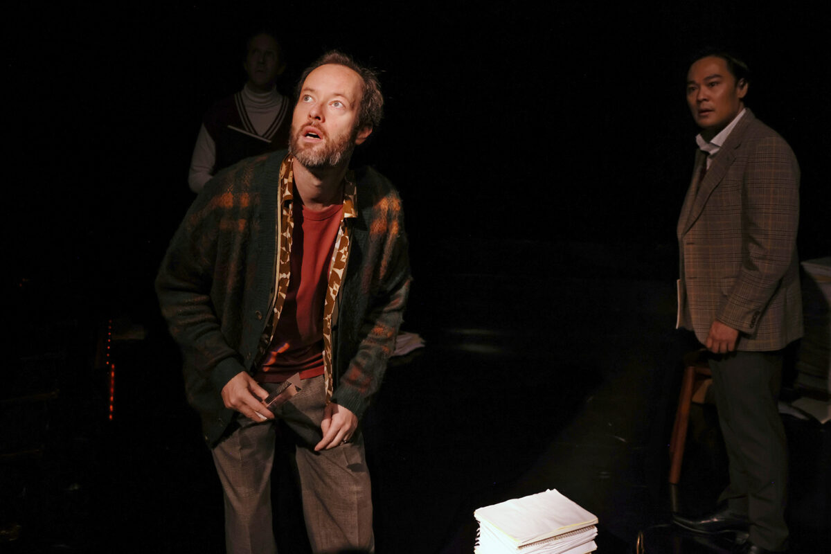 Three men stand in a dimly lit room. The man at the front looks upward with a startled expression, gripping his trousers. Behind him, two men watch, one in a checked jacket near a desk with stacked papers and another in a jumper vest.