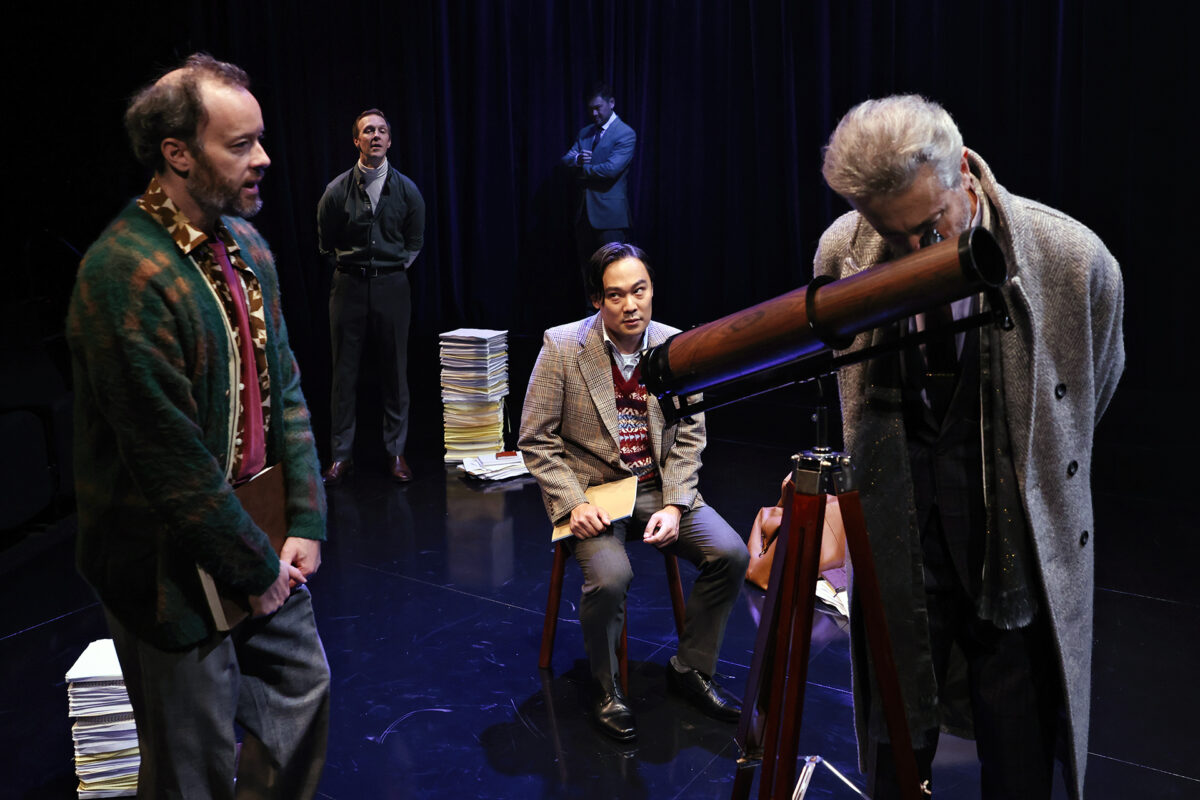 Five men in vintage-style clothing are on a dark stage, surrounded by stacks of papers. One man looks through a telescope whilst the others watch, with expressions of curiosity and contemplation.