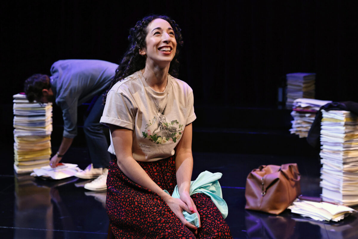 A woman with curly hair sits smiling onstage, wearing a light T-shirt and patterned skirt, holding a light blue cloth. Behind her, a person bends over stacks of papers in a dimly lit setting with a brown bag nearby.