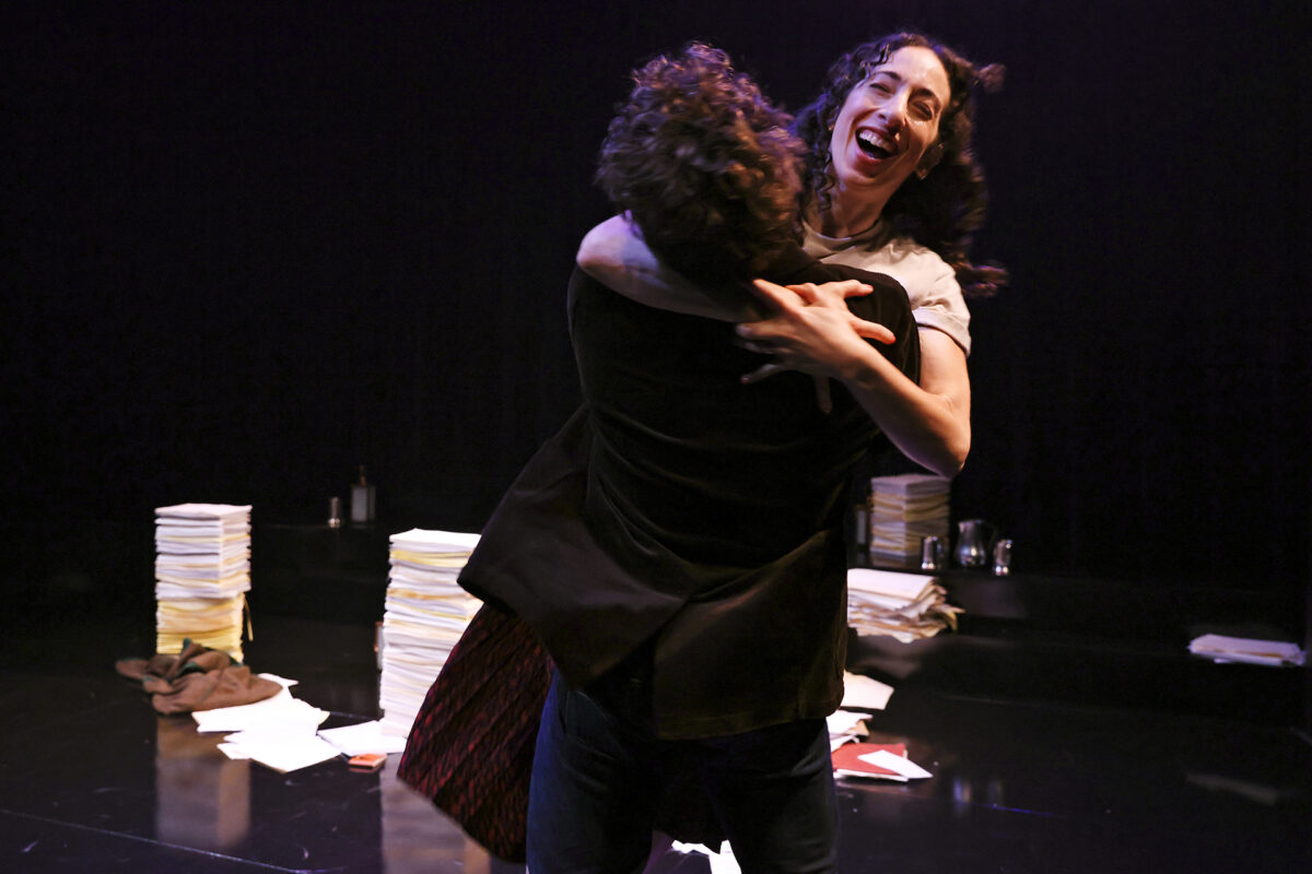 A person with curly hair lifts and embraces a smiling woman on a stage, surrounded by scattered papers and stacks of documents, against a dark background.