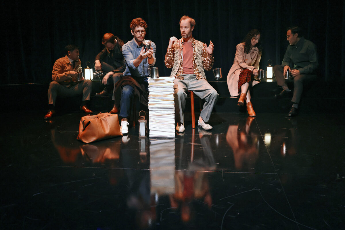 Six people sit on stage in a dimly lit setting, with two men at the centre interacting over a tall stack of papers; others are seated in the background, each with a lantern, suggesting a theatrical performance.