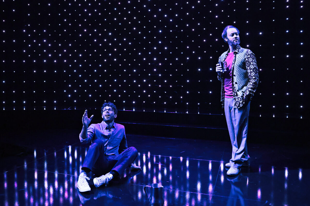 Two men on a stage with a starry lights backdrop; one sits on the floor gesturing upwards, while the other stands holding a microphone, wearing a patterned jacket and gazing into the distance.