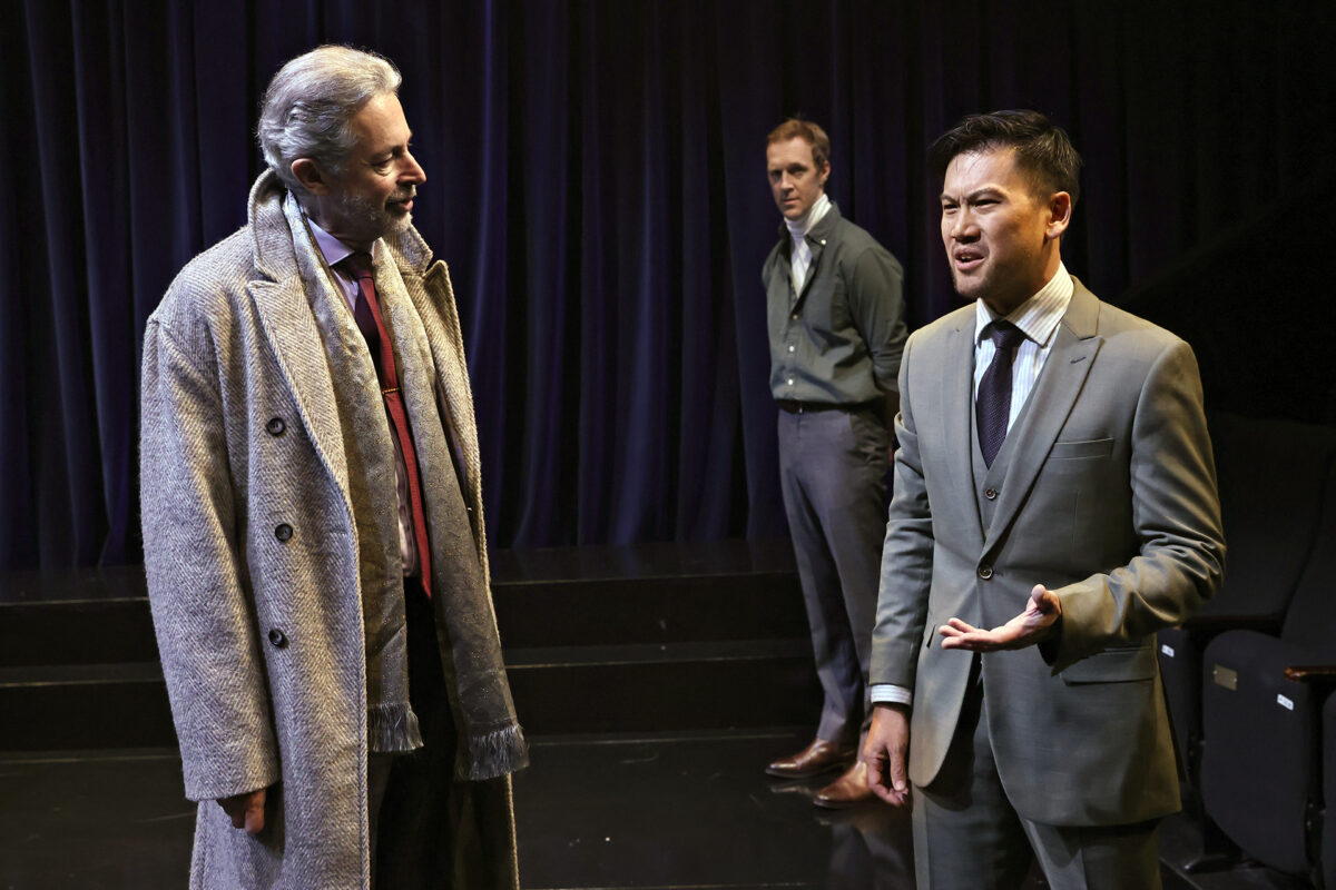 Three men are on stage. One wears a grey overcoat and scarf, another in back wears a cardigan and dress trousers, and the third, in front, wears a light grey suit and gestures while speaking. A dark curtain hangs behind them.