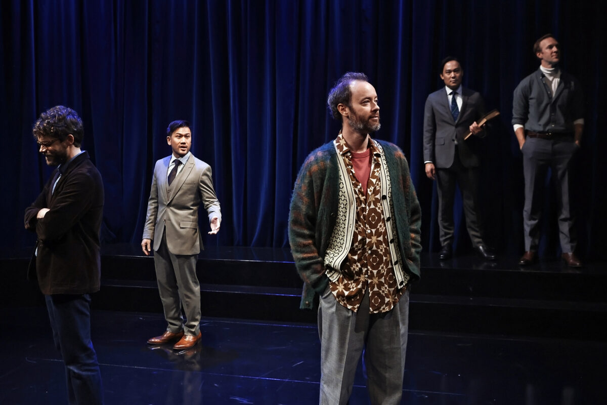 Five men stand on a dimly lit stage with dark blue curtains, dressed in various styles from casual to formal. One stands in front wearing a cardigan, while others stand behind him in suits, each with different poses and expressions.