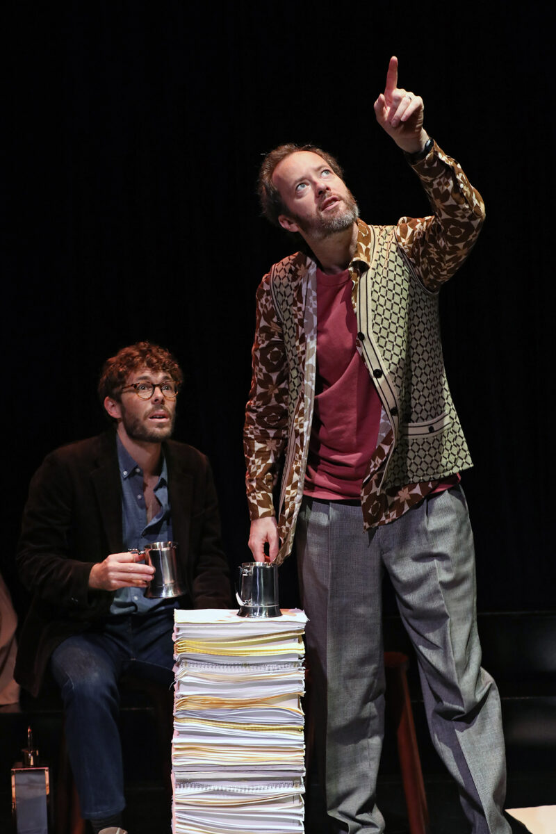Two men on stage, one seated holding a mug and looking up, the other standing beside a tall stack of papers, pointing upward with a focused expression. Both are dressed in casual, eclectic clothing against a dark backdrop.