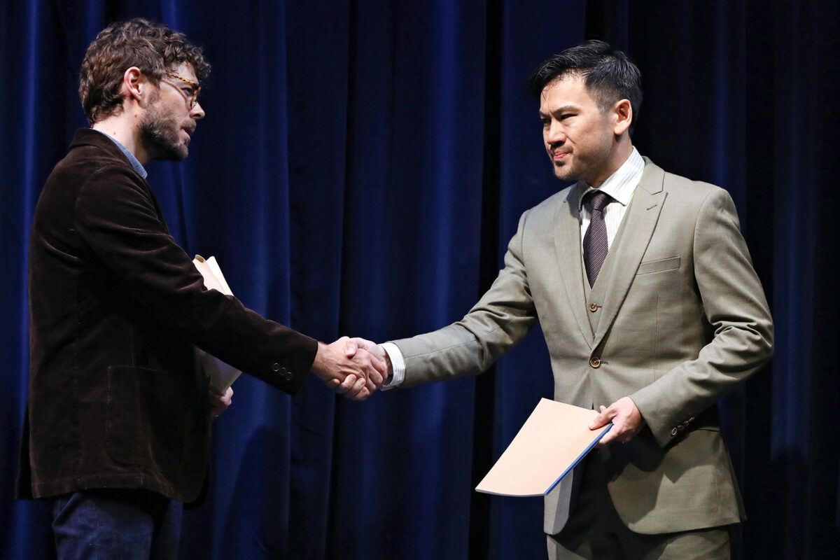 Two men in suits shake hands on stage in front of dark blue curtains. Both hold folders and appear to be engaged in a formal or professional exchange.