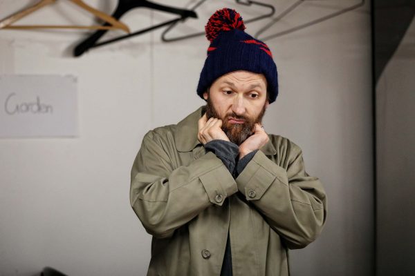 A man in a beige coat and blue winter hat with a red bobble stands indoors, holding his hands to his chin and looking thoughtful. Coat hangers and a sign that says Garden are visible in the background.