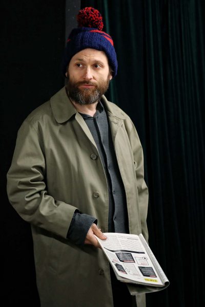 A man with a beard wears a blue and red knitted hat and a green trench coat, holding a printed magazine or newspaper in one hand, standing in front of a dark curtain.