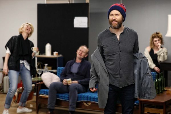 Four adults are in a room; one man at the front puts on a jacket and wears a beanie, while a woman laughs, another man sits on a sofa, and a woman at the back talks on the phone. The setting looks like a casual rehearsal or meeting.