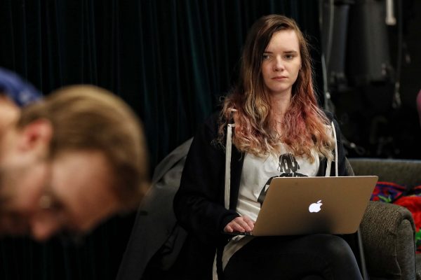 A woman with long, wavy, light brown and pink hair is sitting on a sofa, using a silver MacBook laptop. She is wearing a white shirt and a black hoodie. A blurred person is in the foreground.