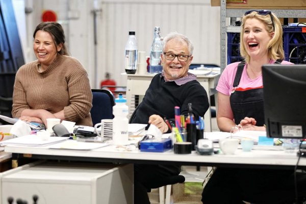 Three people sit at a cluttered desk, laughing and smiling. The group includes two women and one man, surrounded by papers, cups, and water bottles in a brightly lit room with shelves and office supplies.