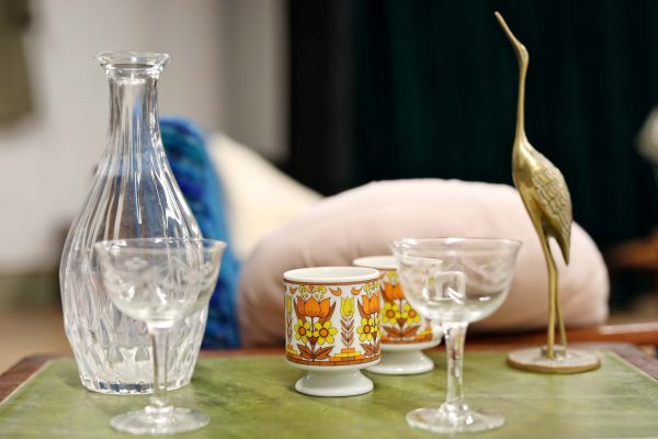 A glass decanter, two empty cocktail glasses, two vintage mugs with floral designs, and a brass crane figurine are arranged on a green surface, with cushions and blurred furniture in the background.