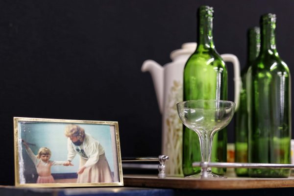 A framed photo of a woman and child sits on a table beside empty green wine bottles, a white teapot with a floral design, a wine glass, and a serving tray, all set against a dark background.
