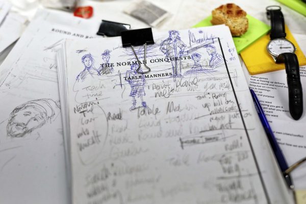 A cluttered desk with a handwritten and doodled script titled The Norman Conquests: Table Manners, surrounded by papers, a pen, a wristwatch, a bulldog clip, a notepad, and a partially eaten piece of bread.