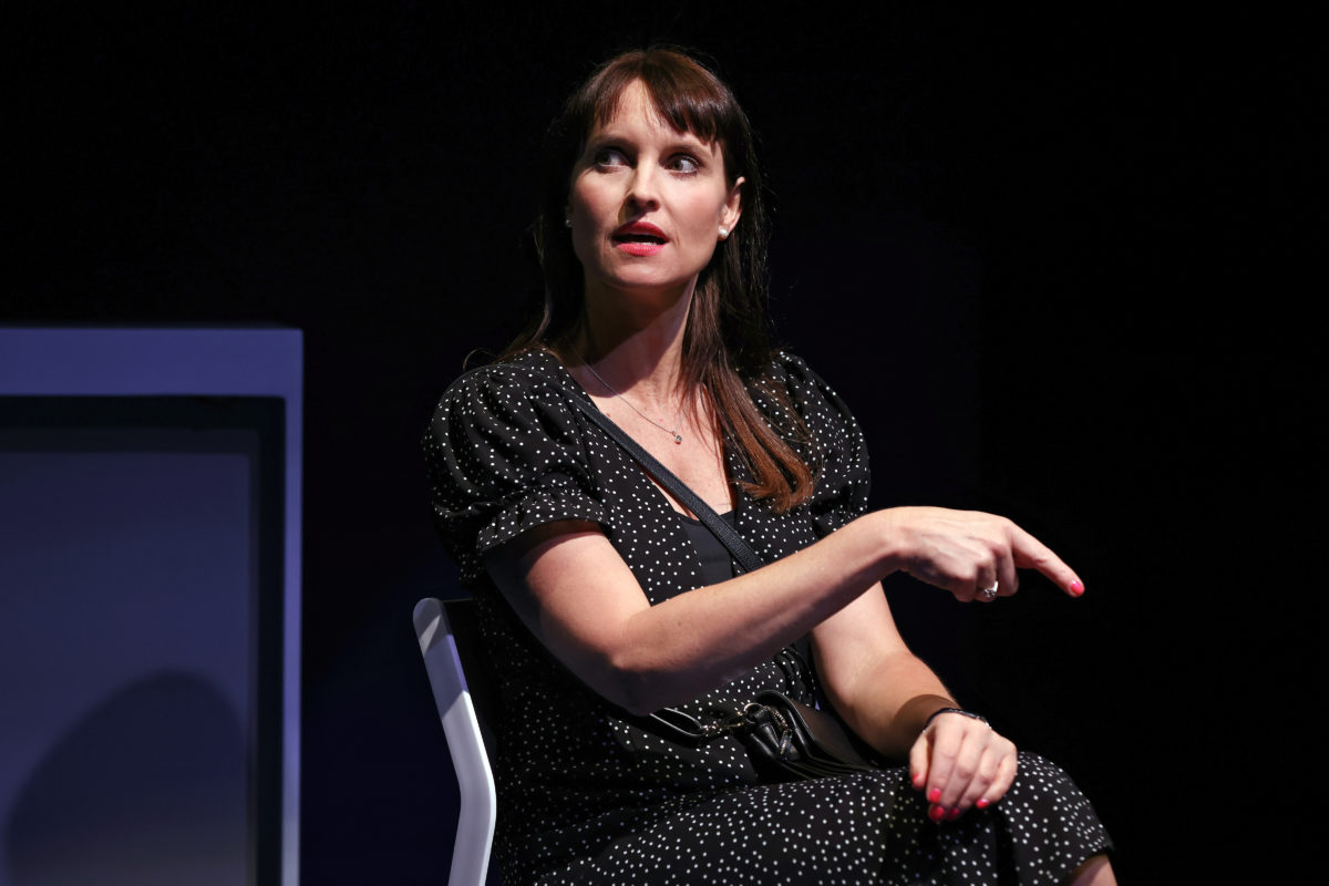 A woman with straight brown hair wearing a black dress with white polka dots sits on a chair against a dark background, looking to her left and gesturing with her right hand, as if speaking or making a point.