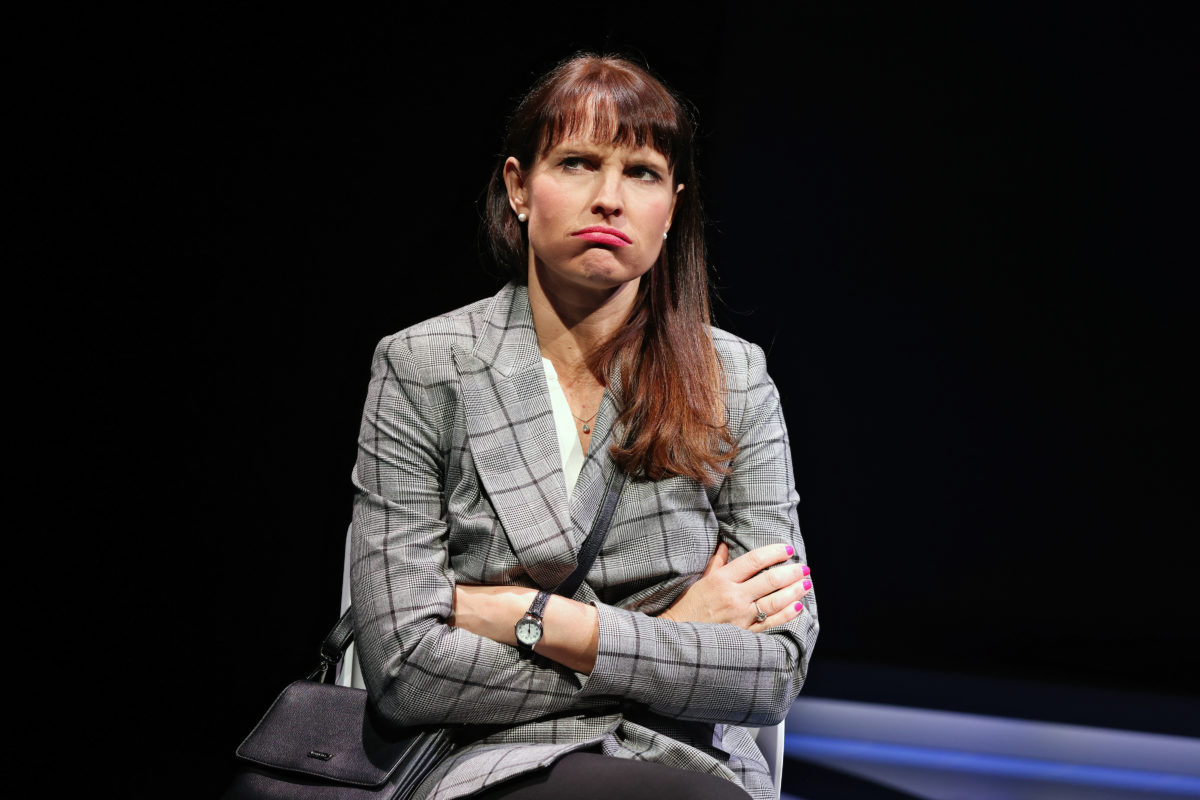 A woman with long brown hair in a checked blazer sits with arms crossed, frowning and looking displeased. The background is dark.