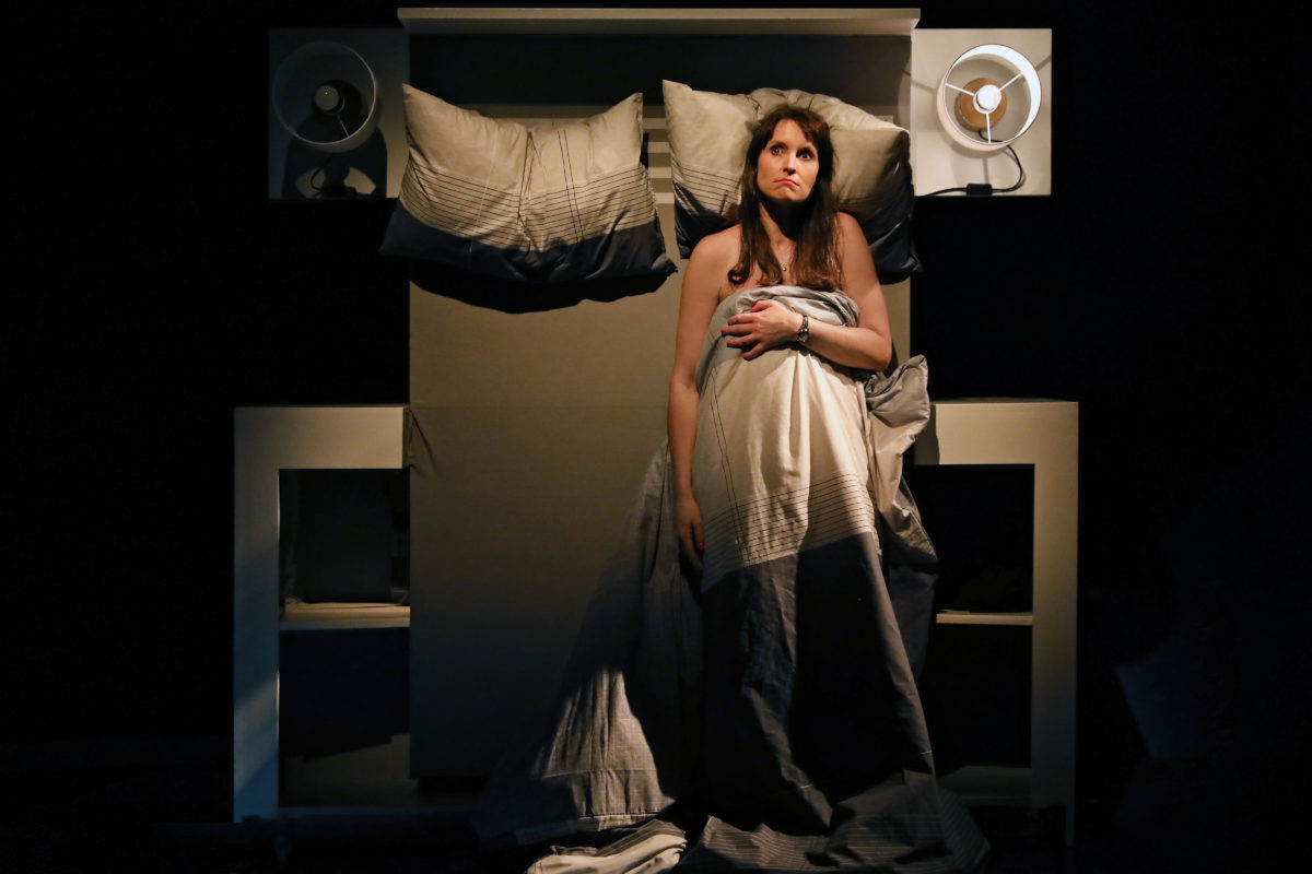 A woman sits upright in bed under a grey-striped blanket, looking upward with a thoughtful expression. The bed is neatly made, flanked by two lamps, with a dark background highlighting the scene.