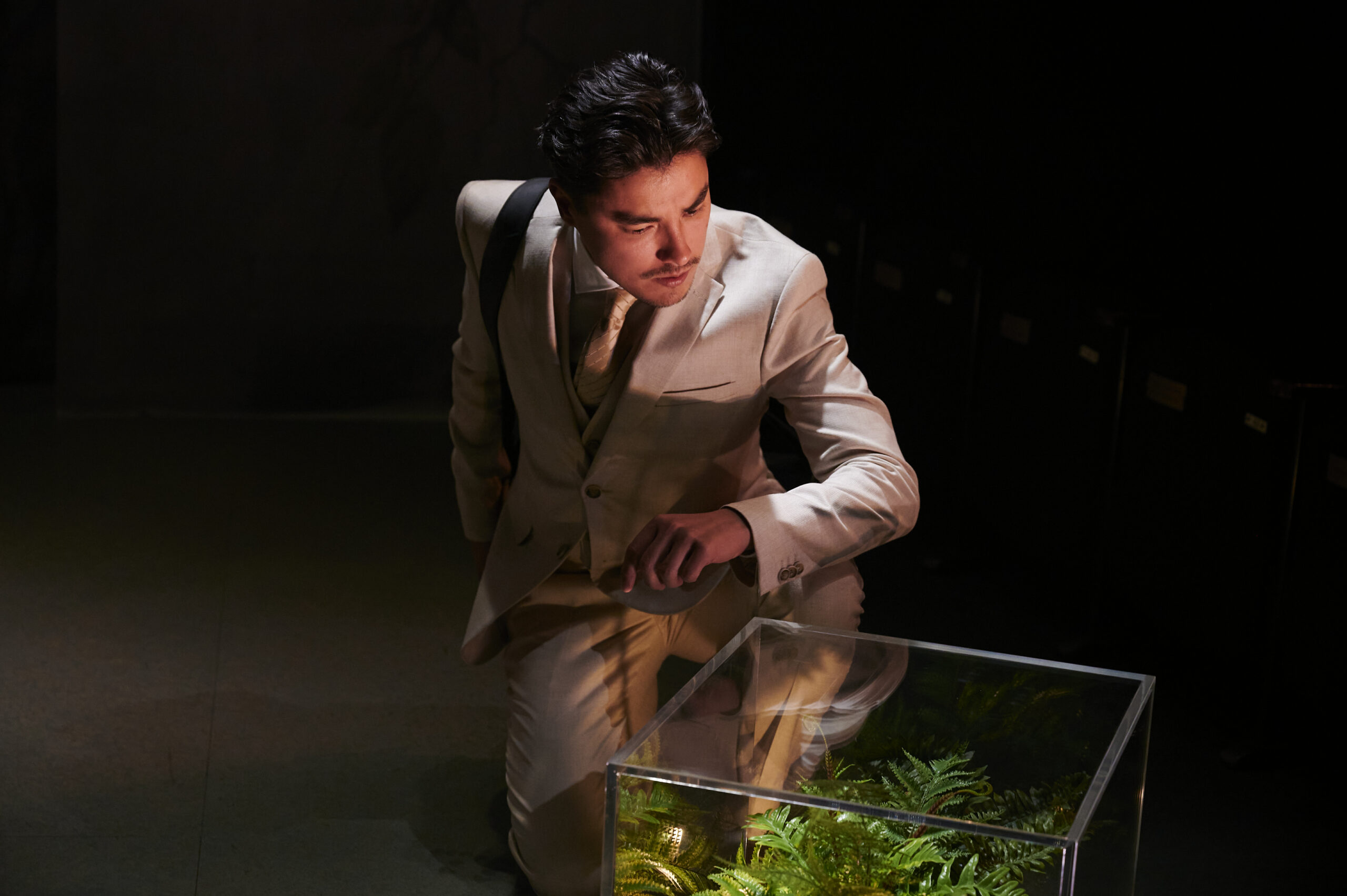 A man in a beige suit kneels under dramatic lighting, looking intently at a glass case filled with green plants. Shadows surround him, creating a focused and contemplative atmosphere.