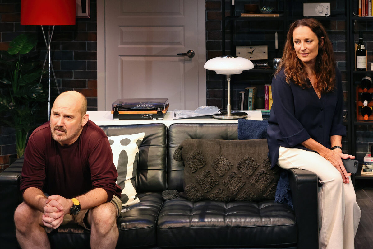 A man and a woman sit on opposite ends of a black sofa in a living room, looking away from each other. The man appears tense, leaning forward, while the woman sits back with a distant expression.