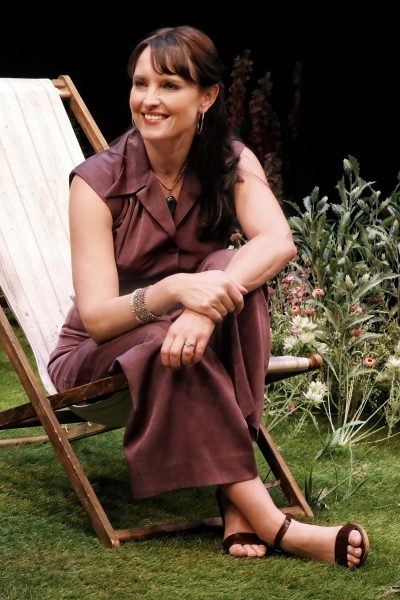 A woman in a mauve jumpsuit sits smiling on a deckchair in a garden setting, surrounded by green grass and flowers.